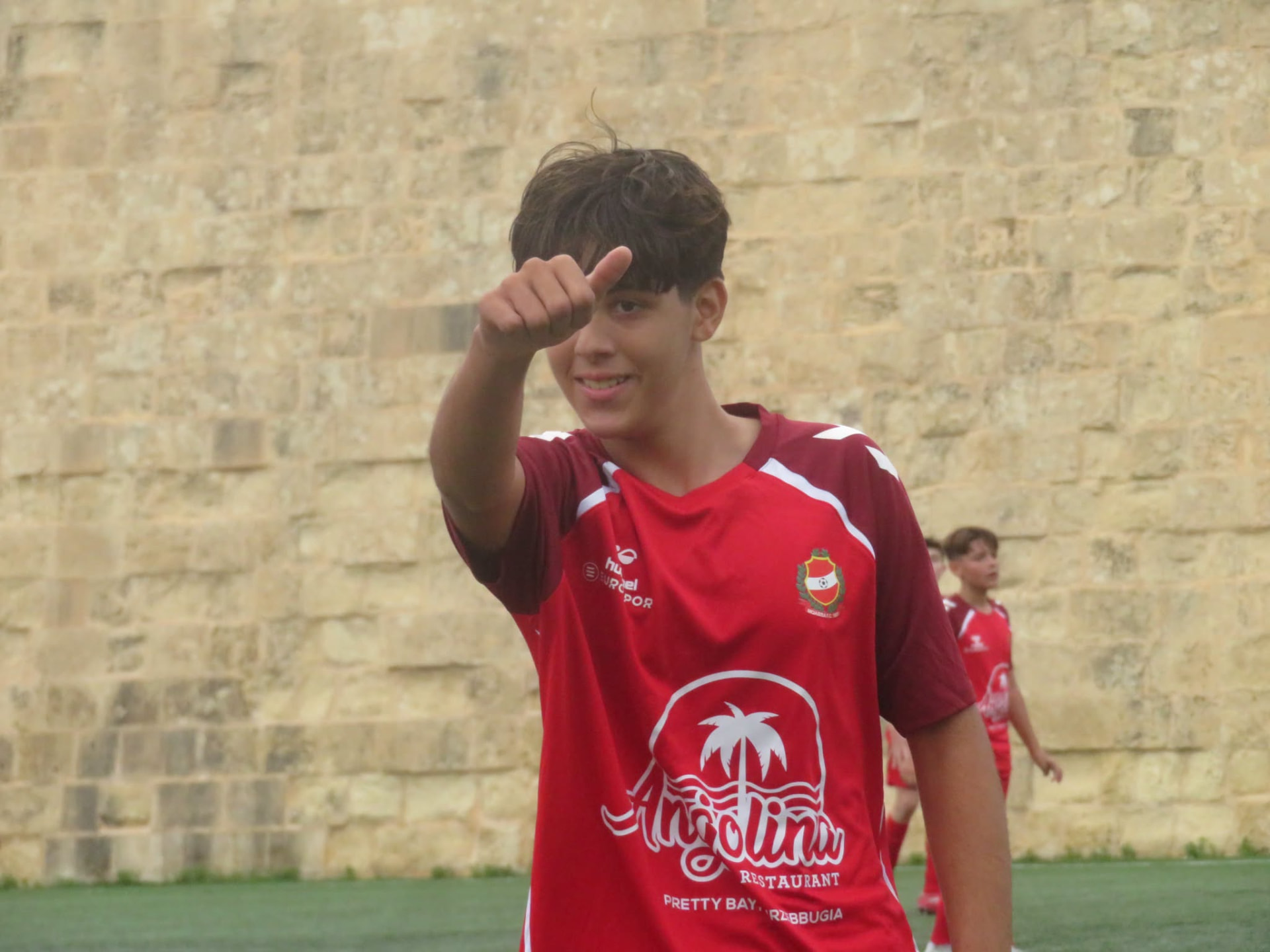 Plejer żagħżugħ bil-flokk aħmar ta' Mqabba F.C. jitbissem u jagħmel thumbs up.