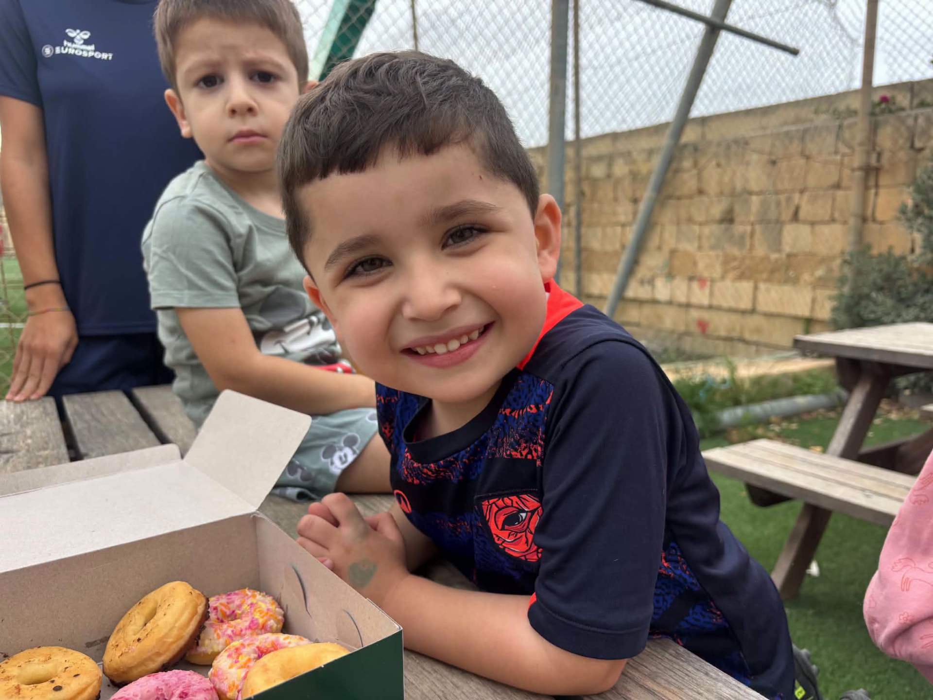 Tifel żgħir jitbissem ħdejn kaxxa doughnuts fuq mejda tal-injam ħdejn il-grawnd.